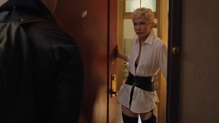 Woman standing in doorway, wearing a white shirt and black belt, with a serious expression.