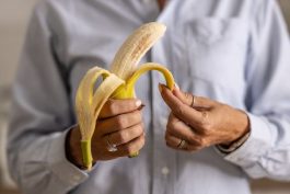 Close-up of a woman's hands peeling a banana. She wears a light blue button-up shirt and rings.